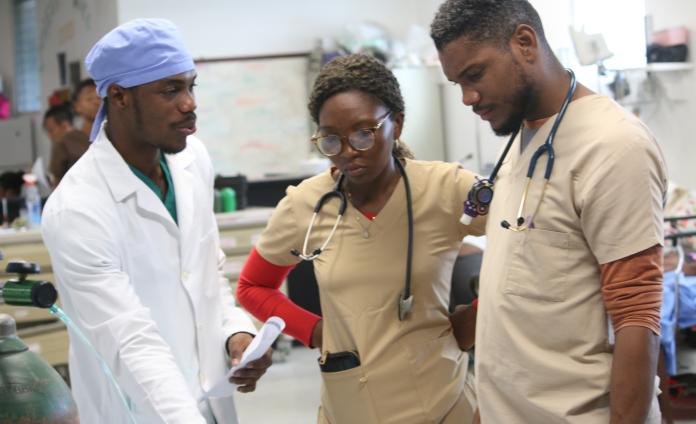 Three clinicians are engaged in a discussion. On the left is Haitian surgeon in a white doctor's coat and a blue surgical cap. In the middle is a woman clinician in braided hair wearing tan scrubs and a stethoscope. On the right is a male Haitian clinician wearing tan scrubs and a stethoscope. 