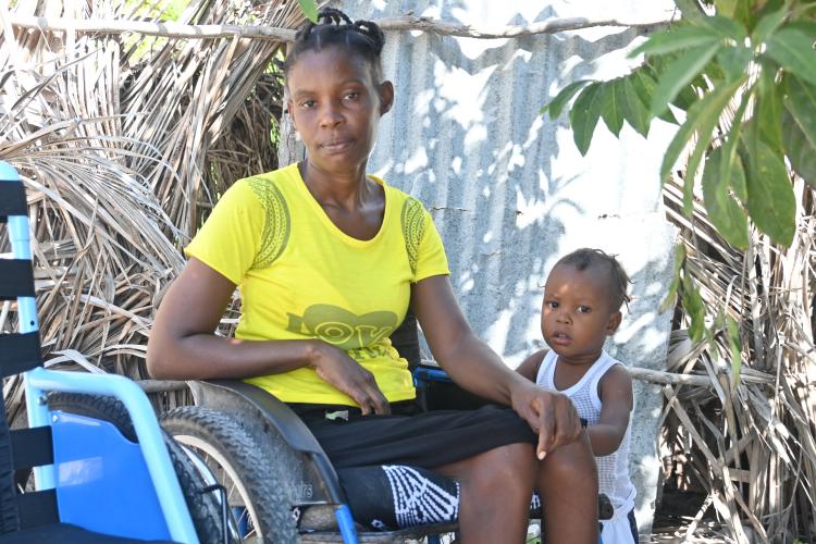 Portrait of a Haitian woman and toddler gazing at the camera. The woman wears a yellow T-shirt and sits in a wheelchair, and the toddler stands close, resting his hand on her leg.