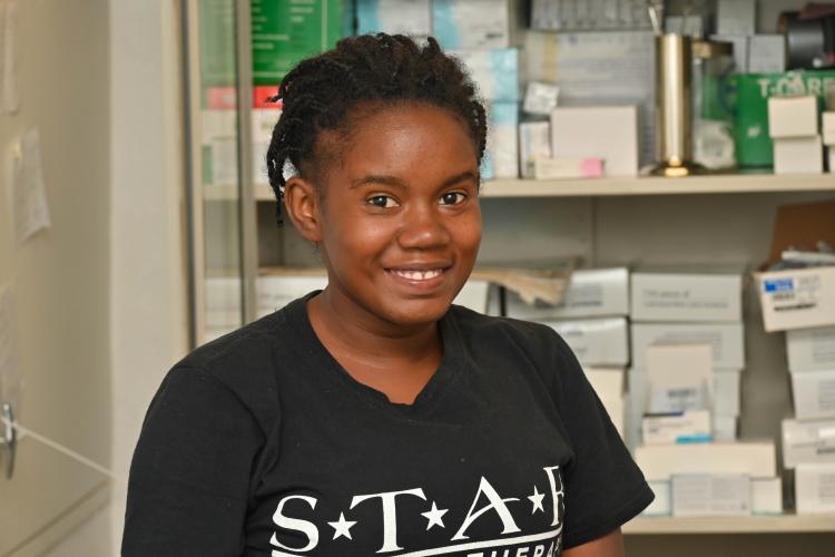 Portrait of a Haitian woman with dimples smiling brightly in front of shelves holding medications. She has short braids and wears a black T-shirt.